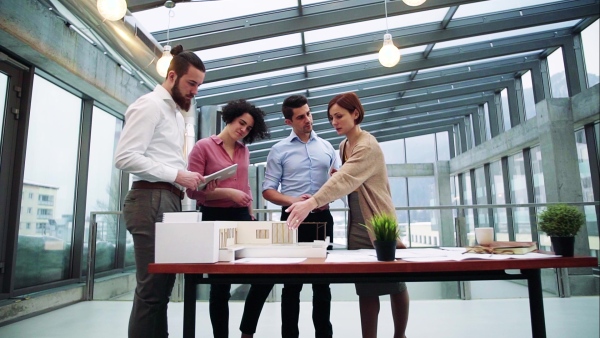 A group of young architects with tablet and model of a house standing in office, working and talking. Slow motion.