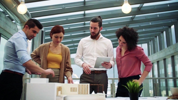 A group of young architects with tablet and model of a house standing in office, working and talking. Slow motion.