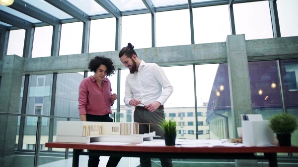 Two young architects with model of a house standing in office, working and talking. Slow motion.