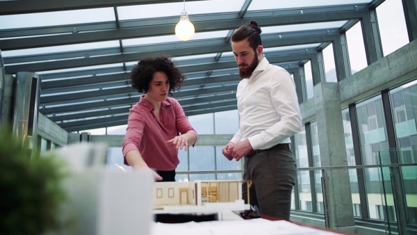 Two young architects with model of a house standing in office, working and talking. Slow motion.