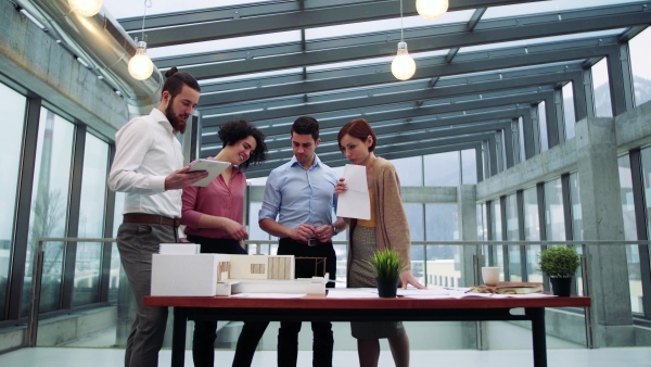 A group of young architects with model of a house standing in office, working and talking.