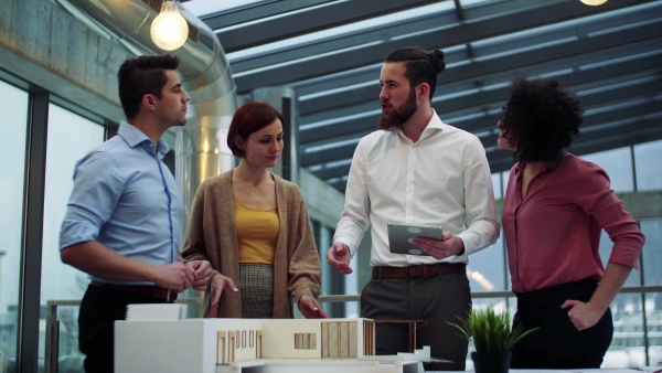 A group of young architects with tablet and model of a house standing in office, working and talking.