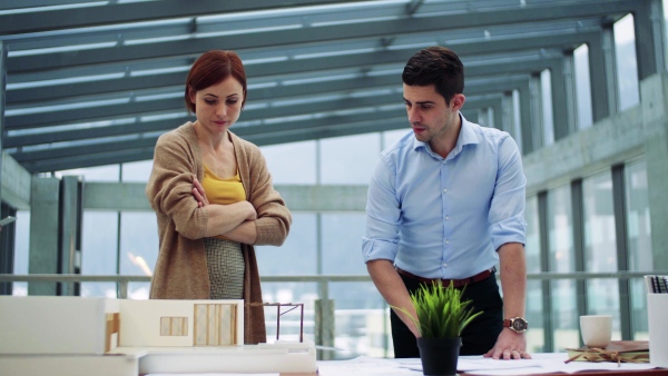 Two young architects with model of a house standing in office, working and talking.