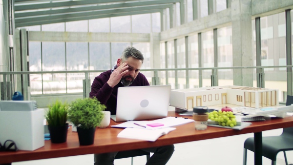 A mature architect with laptop and model of a house in office, working.