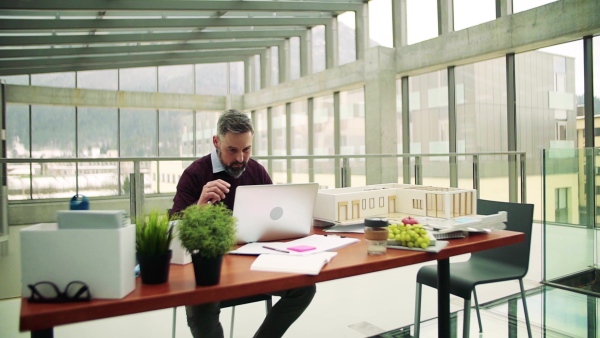 A mature architect with laptop and model of a house in office, working. Slow motion.