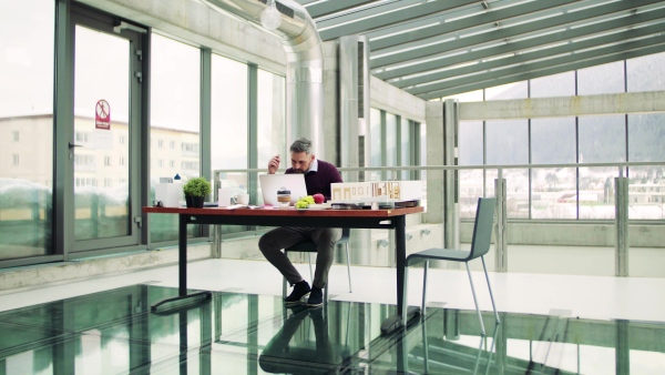 A mature architect with laptop and model of a house in office, working.