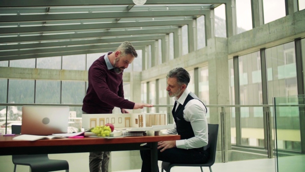 Two male architects with model of a house standing in office, working and talking. Slow motion.