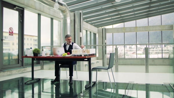 A mature architect with laptop and model of a house in office, working.