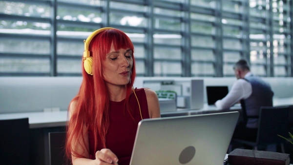 A mature woman with laptop and headphones in office, listening to music. Slow motion.