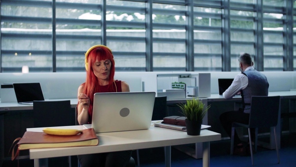 A mature woman with laptop and headphones in office, listening to music. Slow motion.