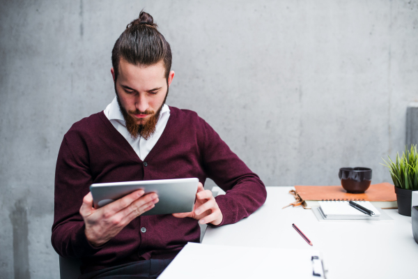 A young businessman with tablet sitting at the desk in office, working.