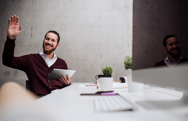 A young businessman with tablet sitting at the desk in office, greeting somebody.