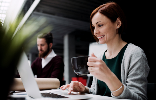 A happy young businesswoman with a cup of coffee working in office, using laptop.