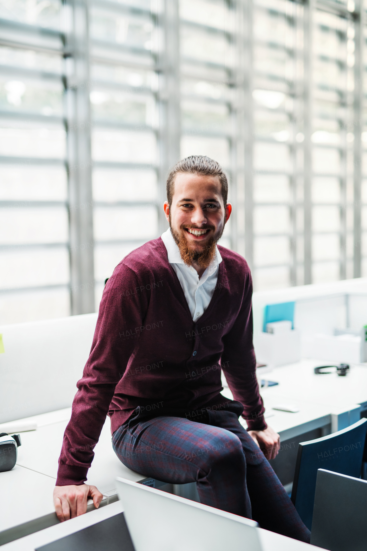 A portrait of cheerful young businessman sitting on a desk in office, looking at camera.