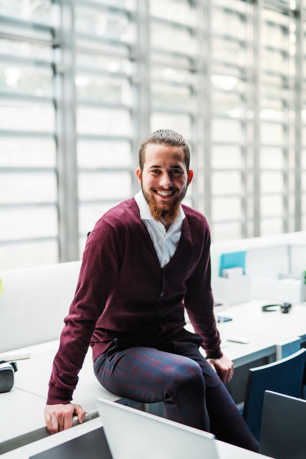 A portrait of cheerful young businessman sitting on a desk in office, looking at camera.