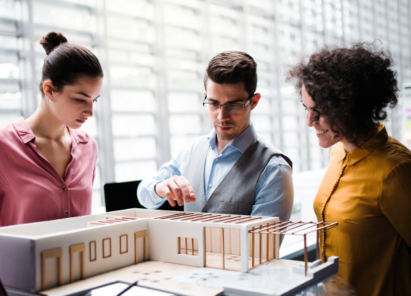 A group of young architects with model of a house standing in office, working and talking.