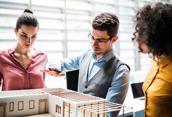 A group of young architects with model of a house standing in office, working and talking.