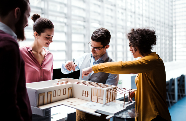 A group of young architects with model of a house standing in office, working and talking.