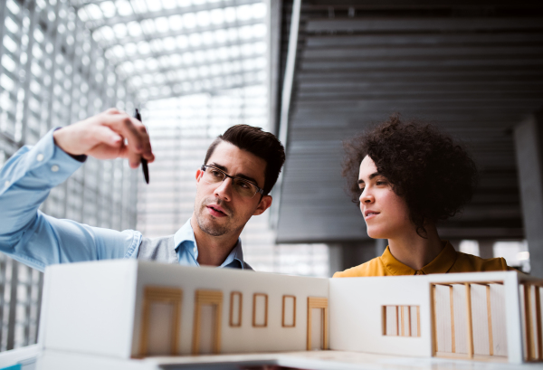 Two young architects with model of a house standing in office, working and talking.