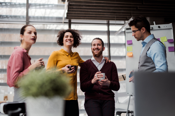 A group of cheerful young businesspeople with cup of coffee standing in office, talking.