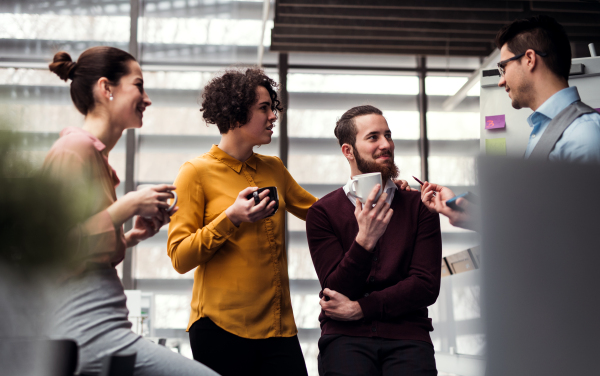 A group of cheerful young businesspeople with cup of coffee standing in office, talking.