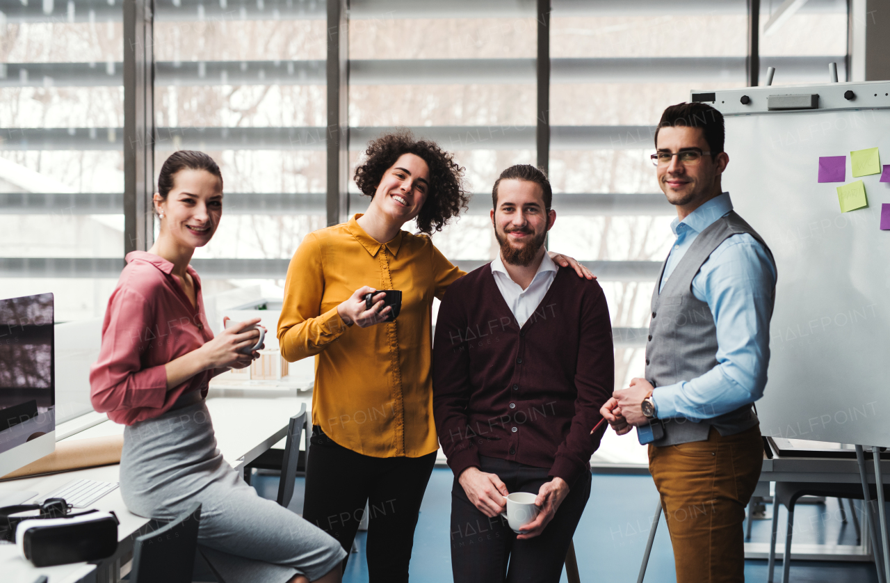 A group of cheerful young businesspeople with cup of coffee standing in office, looking at camera.