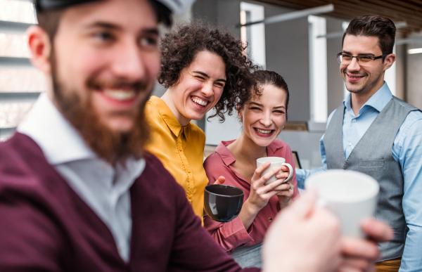 A group of cheerful young businesspeople with cup of coffee standing in office, laughing.