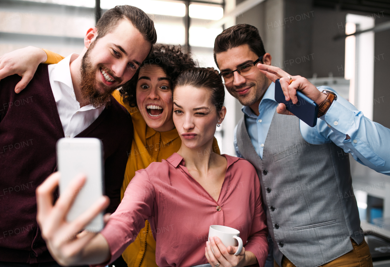 A group of cheerful young businesspeople with smartphone in office, taking selfie.