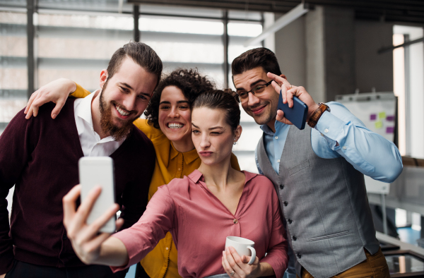 A group of cheerful young businesspeople with smartphone in office, taking selfie.