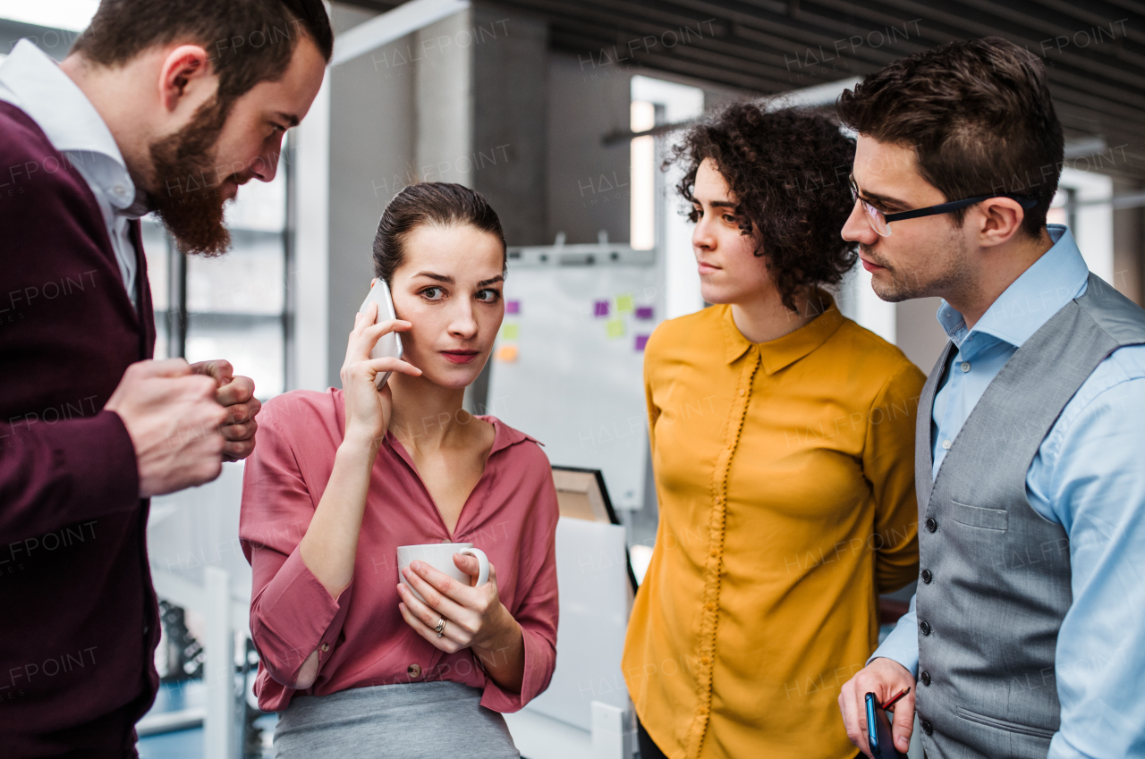 A group of young cheerful businesspeople with smartphone standing in office, making a phone call.