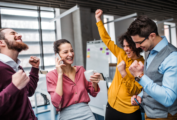 A group of young cheerful businesspeople with smartphone standing in office, expressing excitement.