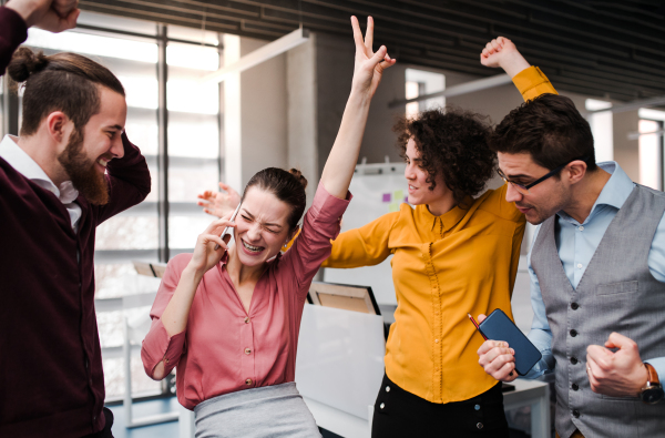 A group of young cheerful businesspeople with smartphone standing in office, expressing excitement.