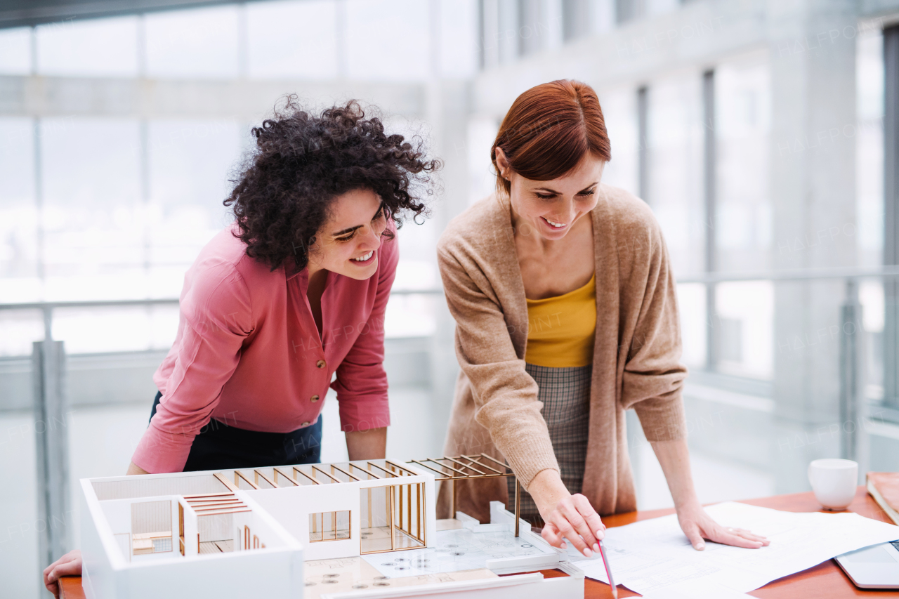 Two female young architects with model of a house standing in office, working and talking.