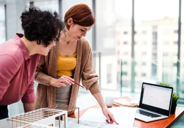 Two female young architects with model of a house standing in office, working and talking.