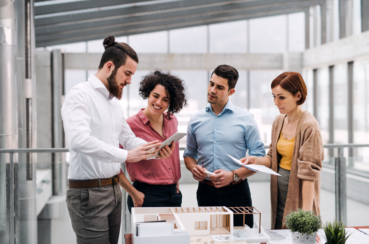 A group of young architects with tablet and model of a house standing in office, working and talking.