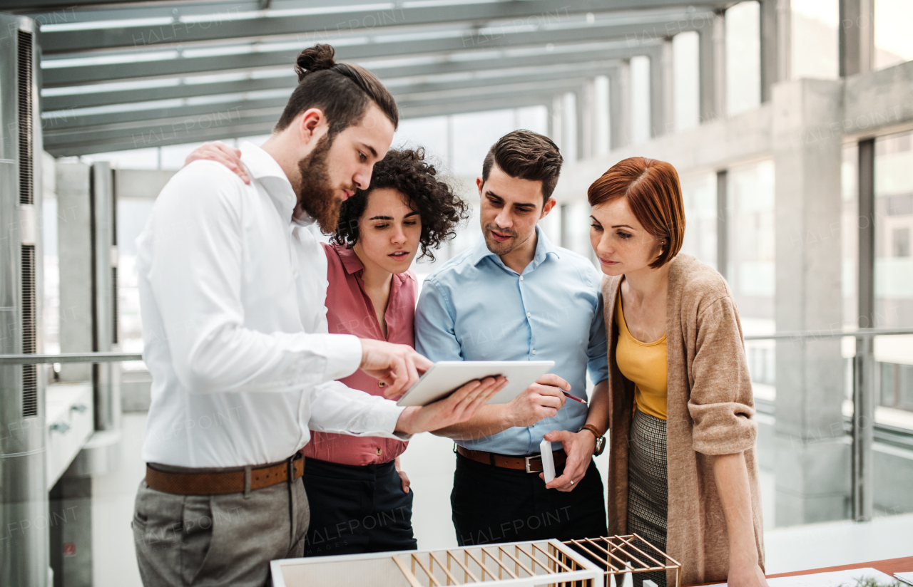 A group of young architects with tablet and model of a house standing in office, working and talking.