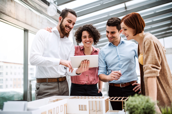 A group of young architects with tablet and model of a house standing in office, working and talking.