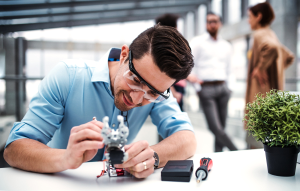 A young businessman or scientist with robotic hand and safety glasses standing in office, working.