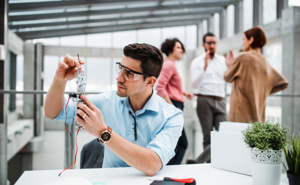 A young businessman or scientist with robotic hand and safety glasses standing in office, working.