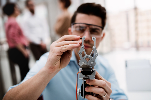 A young businessman or scientist with robotic hand and safety glasses standing in office, working.