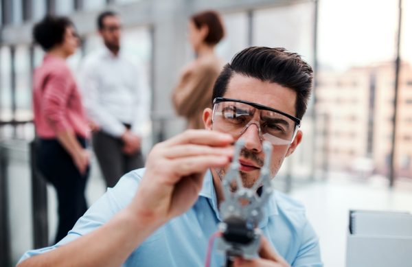 A young businessman or scientist with robotic hand and safety glasses standing in office, working.