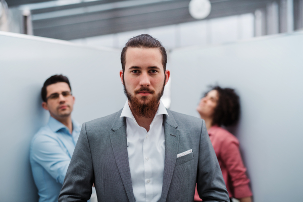 A portrait of young businessman in suit with colleagues standing in office.