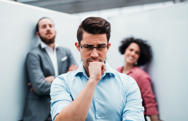 A portrait of young thoughtful businessman standing in office, colleagues in the background.