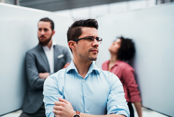 A portrait of young businessmanstanding in office, colleagues in the background.