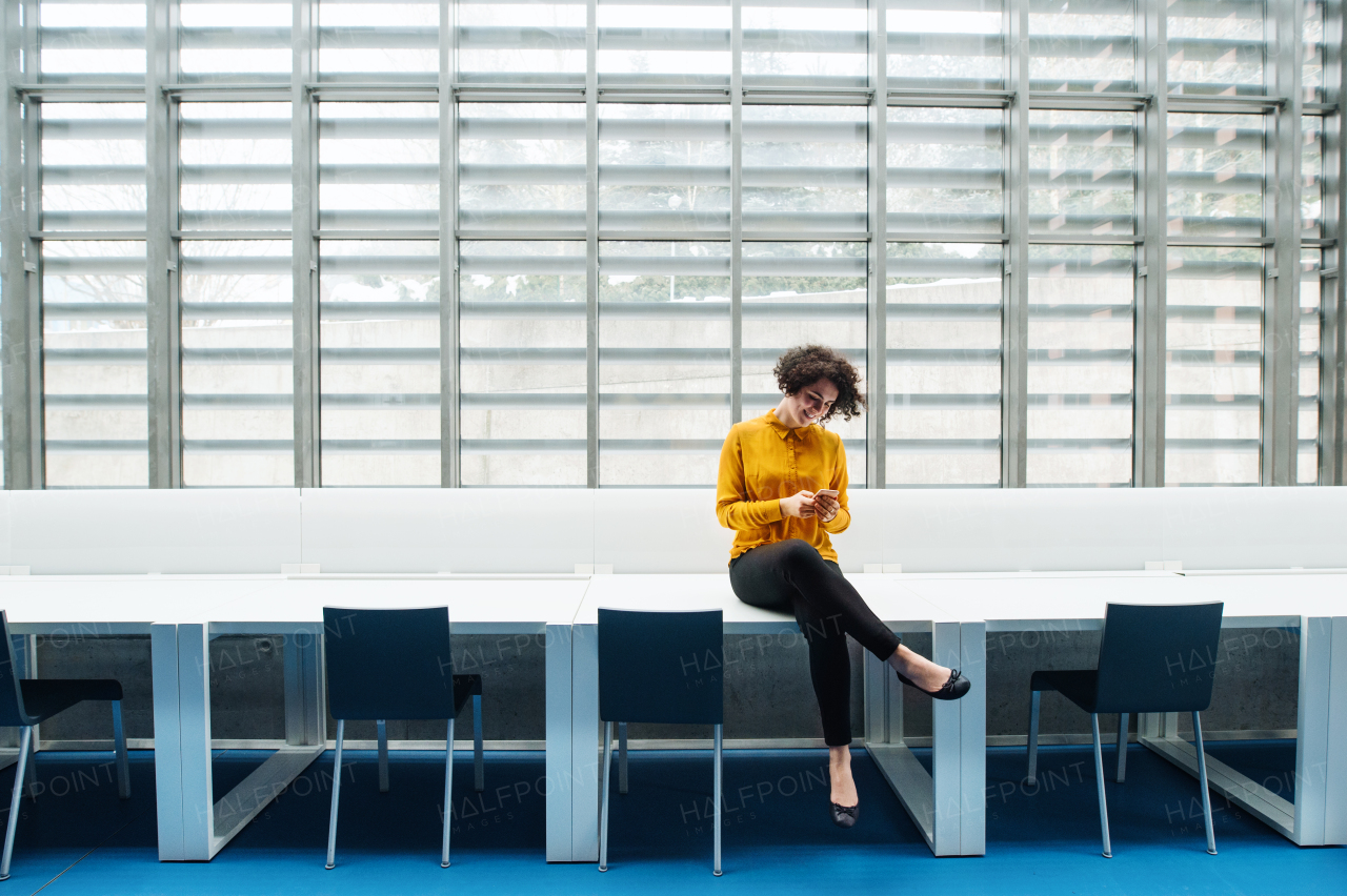 Young student or businesswoman sitting cross-legged on desk in room in a library or office, using smartphone. Copy space.