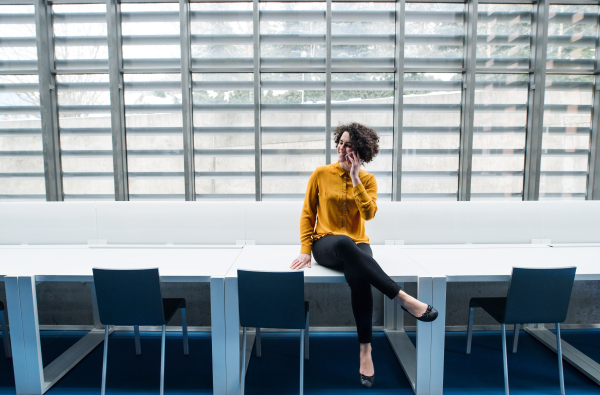 Young student or businesswoman sitting cross-legged on desk in room in a library or office, using smartphone. Copy space.