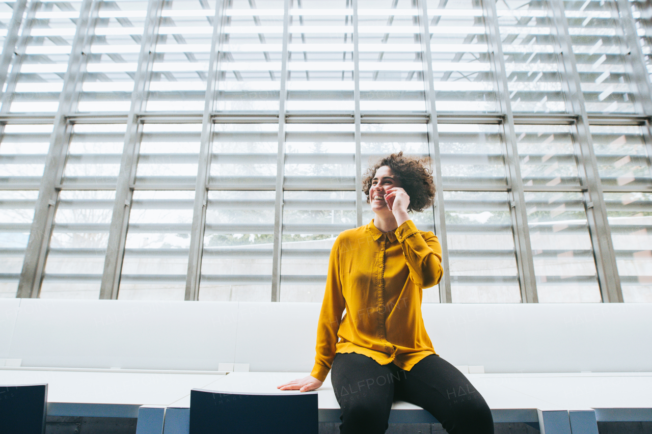 A portrait of a young student or businesswoman sitting on desk in room in a library or office, using smartphone.