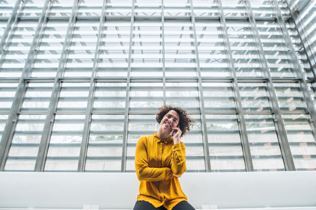 A portrait of a young student or businesswoman sitting on desk in room in a library or office, using smartphone.