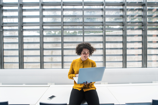 A portrait of a young student or businesswoman sitting on desk in room in a library or office, using laptop.