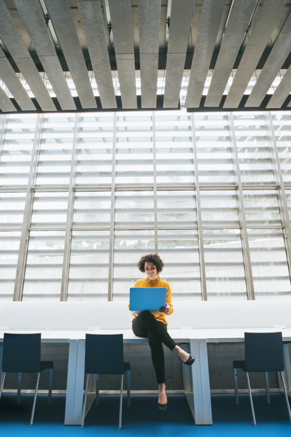 A portrait of a young student or businesswoman sitting on desk in room in a library or office, using laptop.